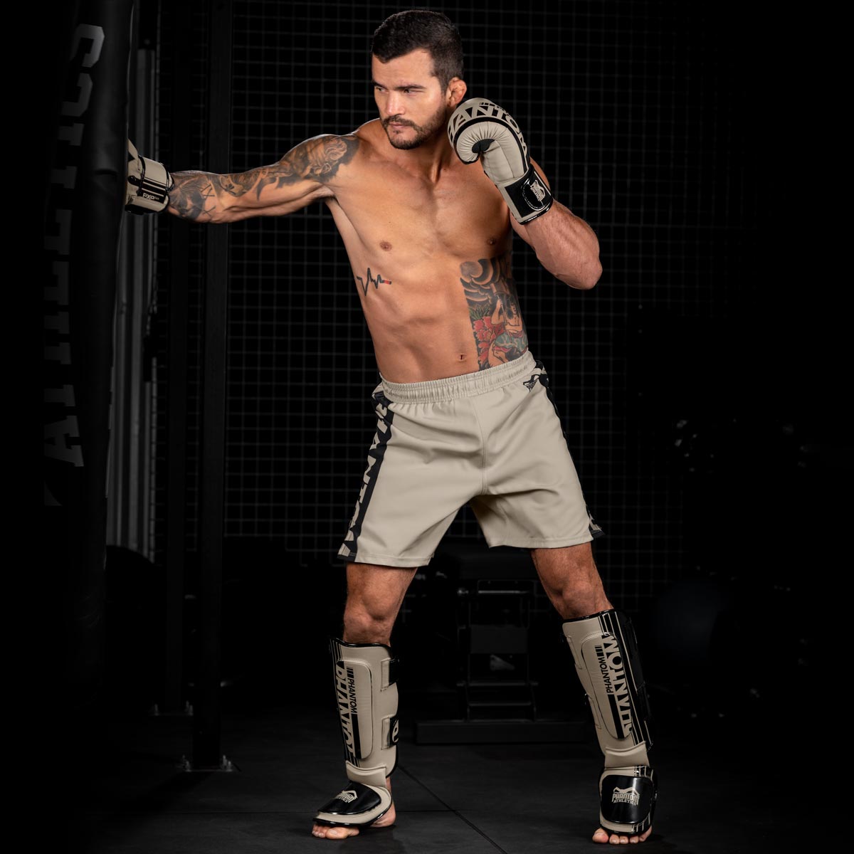 MMA Fighter beim Training am Boxsack mit dem Phantom Apex Fightequipment in Sand. Bestehend aus Phantom Apex Boxhandschuhen, MMA Shorts und Hybrid Schienbeinschonern.